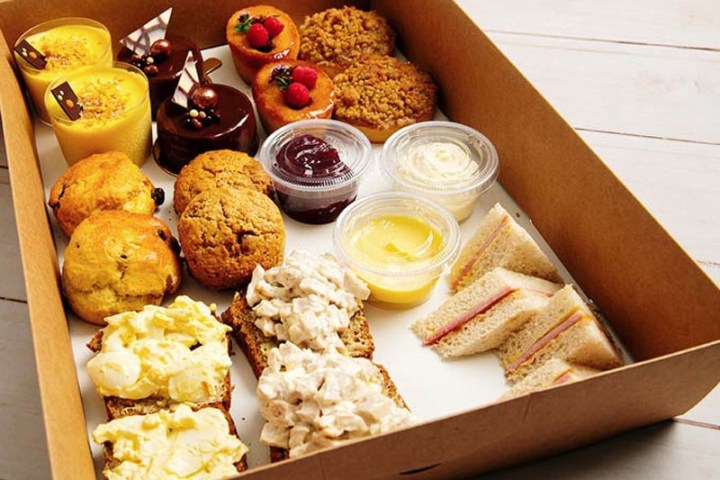Assorted pastries, sandwiches, and condiments in a cardboard box on a wooden surface.