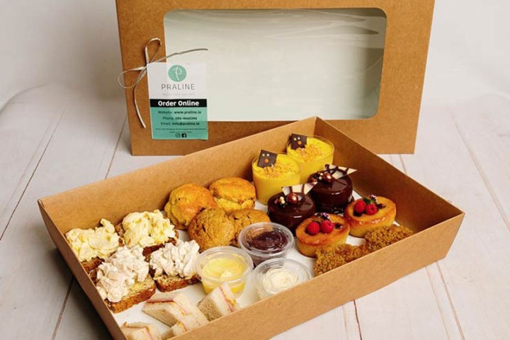 Open food box with sandwiches, scones, pastries, and dips on a wooden surface.