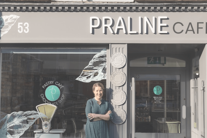 A person stands smiling outside Praline Cafe next to a large window displaying a pastry and chocolate shop sign.