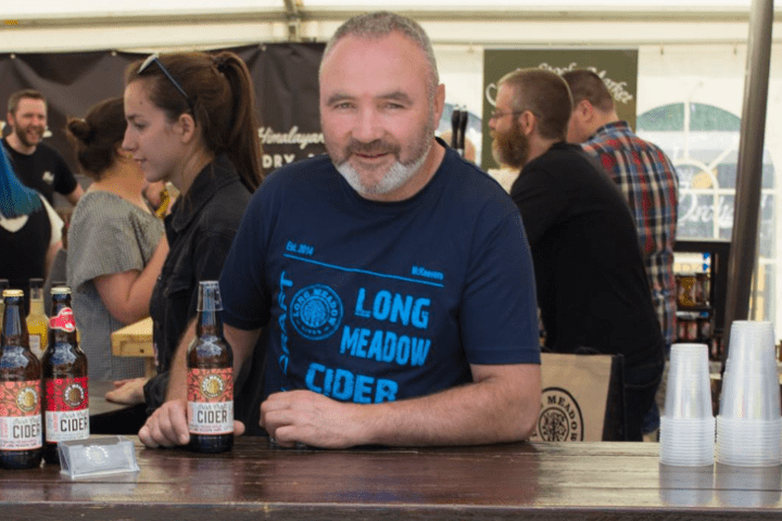 Man in blue shirt standing at a cider booth with bottles and plastic cups on the counter.