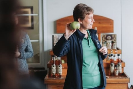 Woman holding an apple, standing in front of a table with bottles in a room.