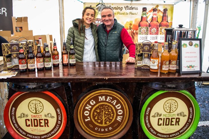Two people behind a cider stall with bottles and signage displaying 'Long Meadow Cider.'