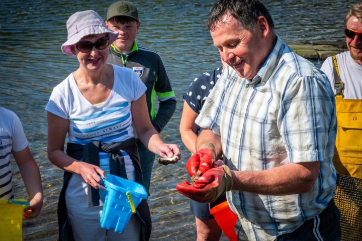 People smiling and gathering shellfish by a river, one man holding a shell.