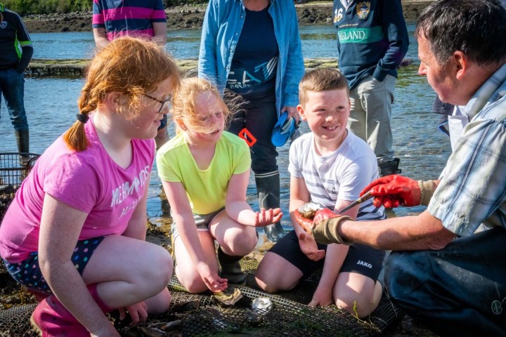Children listen to a man explaining oysters by the water.