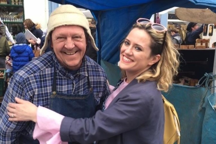 Smiling man and woman in market setting with blue tent and various goods.