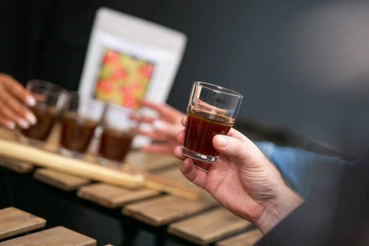 Person holding a small glass of coffee, with more glasses on a tray in the background.