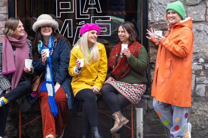 Five women in colorful outfits laughing and holding cups on a stone wall background.