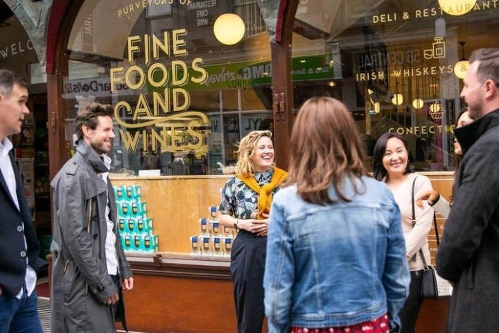 Group of five people chatting outside a deli with 'Fine Foods and Wines' sign.