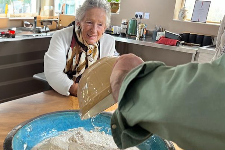 Person pours liquid into a bowl of flour while a smiling woman watches in a kitchen.
