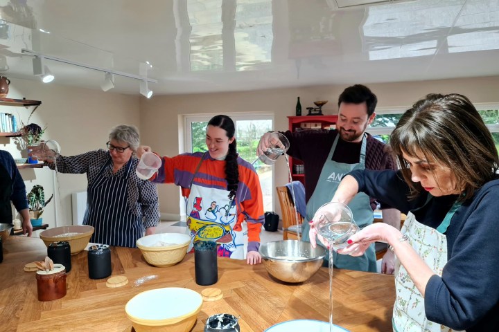 Four people pouring water into bowls at a kitchen counter with focused expressions.
