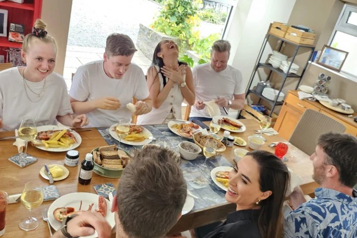 Group of people laughing and eating at a dining table with large windows and bright light.