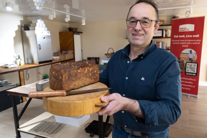 Smiling man holding a large loaf of bread on a wooden cutting board in a kitchen.