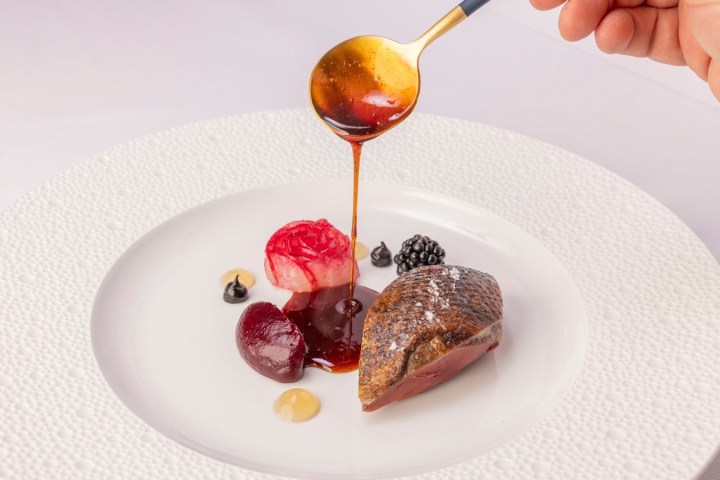 A person drizzles sauce over gourmet food with a spoon on a white plate.