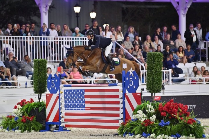 Equestrian jumping over a fence with American flag design, surrounded by spectators.