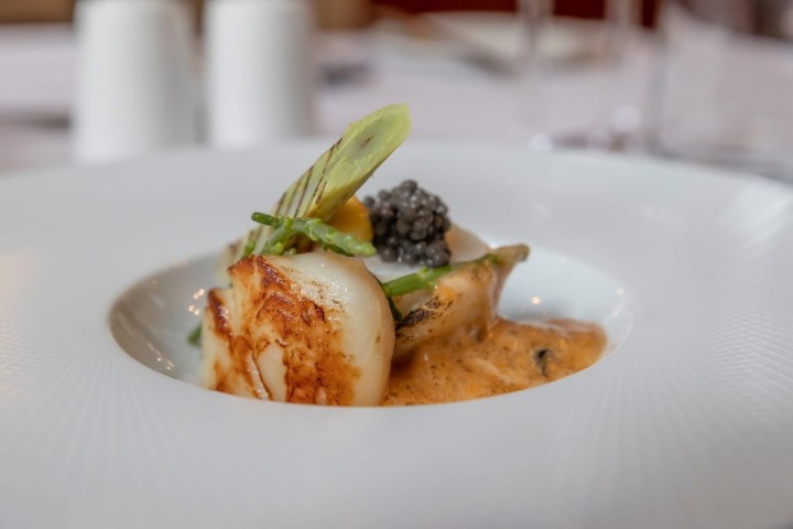 Gourmet dish with a scallop, asparagus, caviar, and sauce on a white plate.