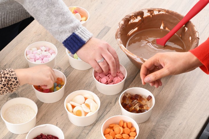 Hands reaching into cups of candies and chocolates on a table with a bowl of melted chocolate.