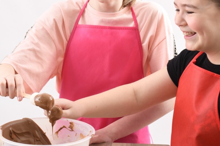Two kids in aprons smiling while mixing chocolate with bowls of candy on the table.