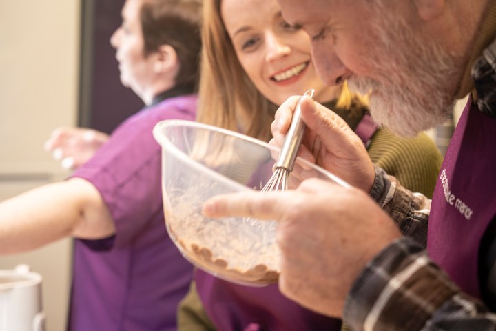 Three people cooking; man whisks ingredients in a bowl while others smile nearby.