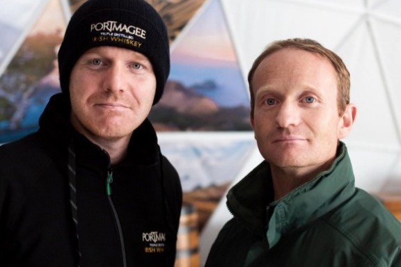 Two men in outdoor jackets stand indoors, one wearing a beanie hat. Triangular decor is visible in the background.