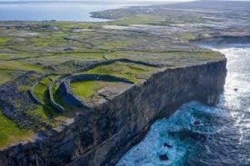 Cliffside landscape with ancient stone fort overlooking ocean and green fields.
