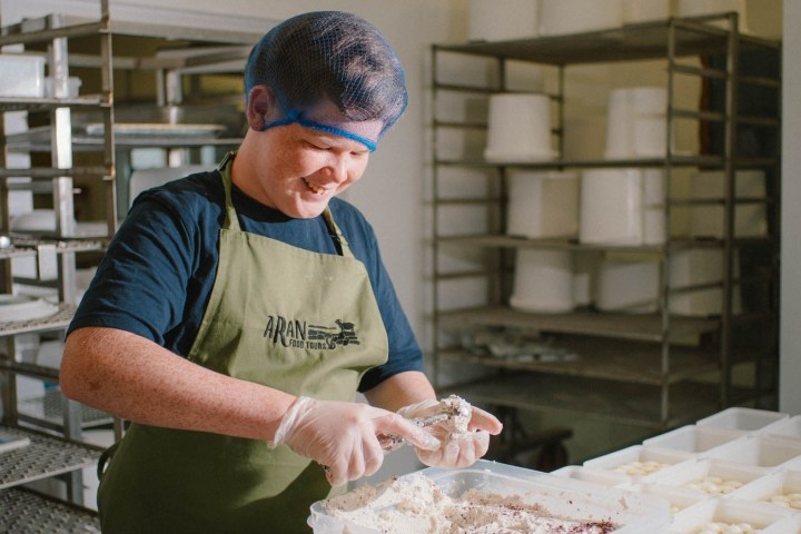 Person in apron and hairnet smiling while handling cheese in a processing room.