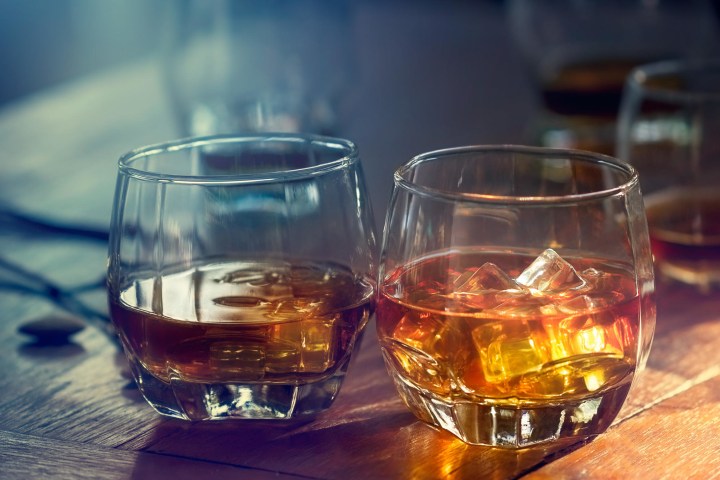 Two glasses of whiskey on a wooden table, one with ice cubes and sunlight filtering through.