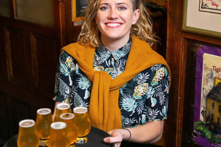 Smiling person holding a tray with six glasses of beer in a pub setting.
