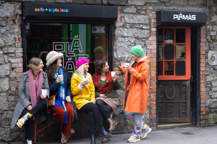 Five people in colorful outfits chatting and holding drinks outside a café with a stone wall.