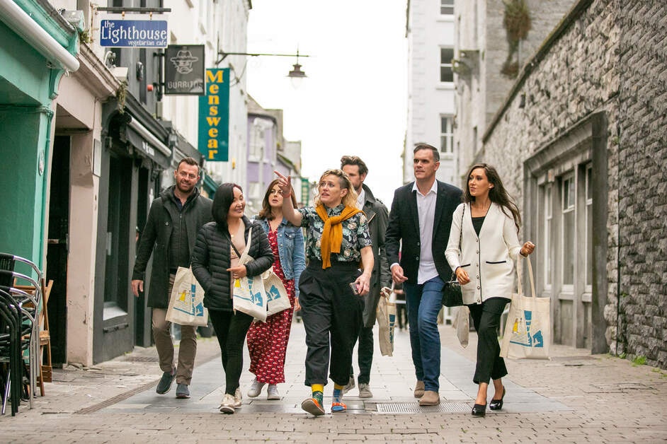 Group of people walking on a narrow street with shops and signs in the background.