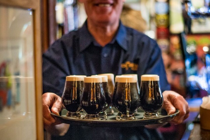 Smiling person holding a tray with six small glasses of dark beer in a cozy pub setting.