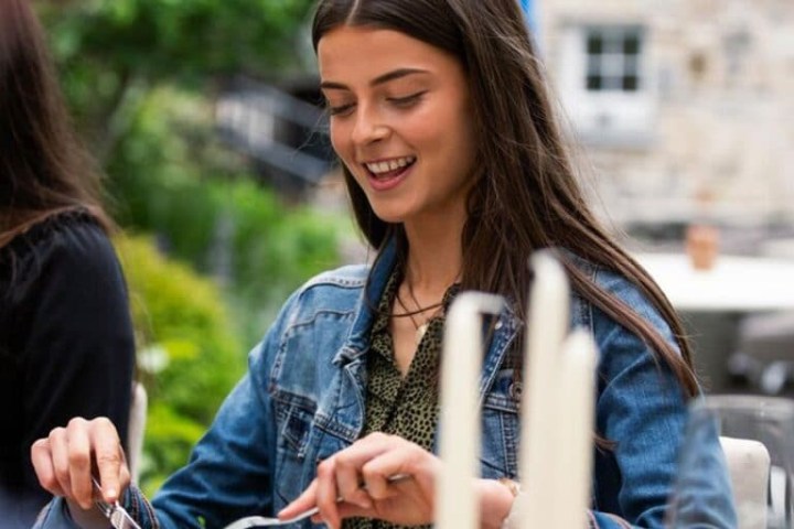 Woman in denim jacket smiling while dining outdoors with salad and drinks.