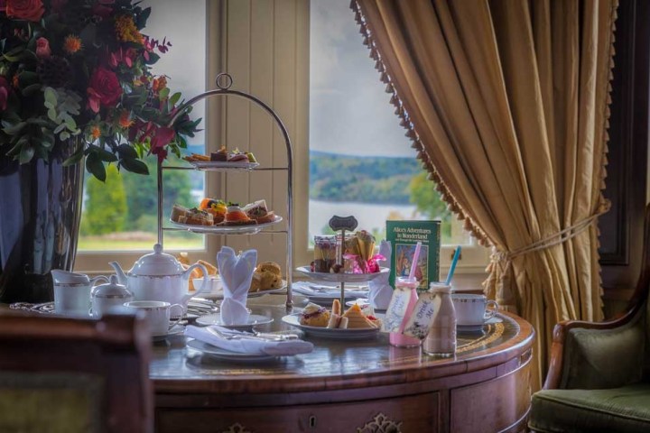 Elegant afternoon tea setup with tiered tray, teapot, and flowers by a window with a lake view.