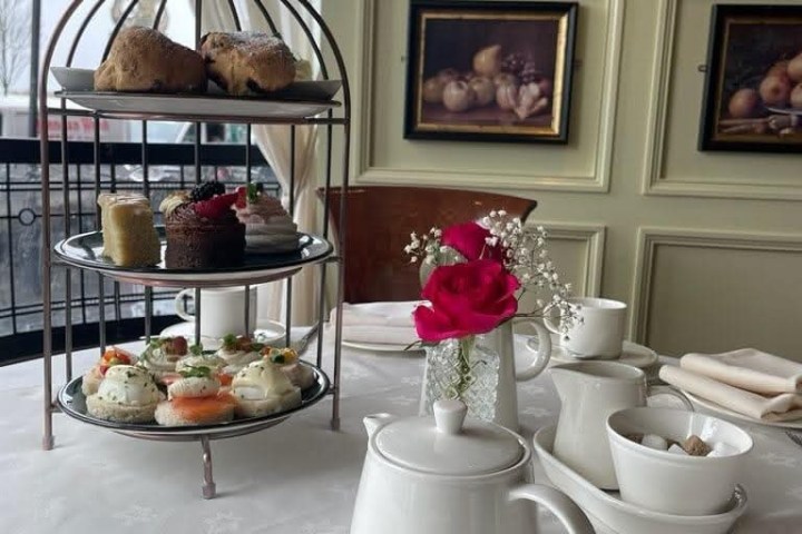 Three-tier tray with snacks, teapot, and pink rose on a table in elegant setting.