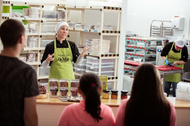 A person in a green apron talks to visitors in a chocolate store kitchen.