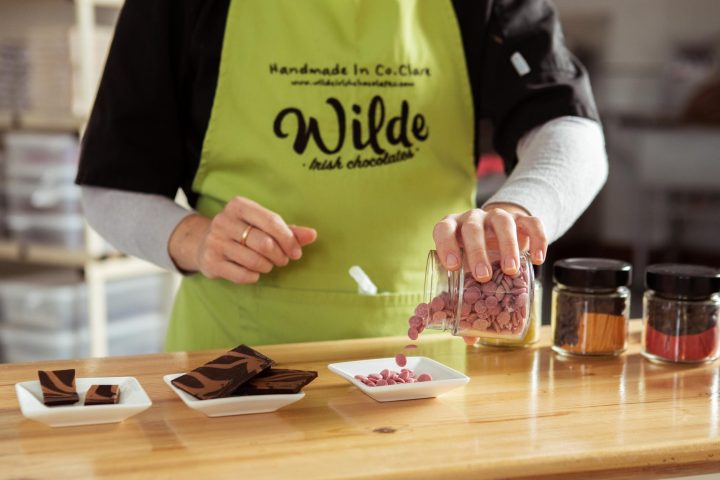 Person in green apron pouring candy into dish beside chocolate squares on a wooden table.