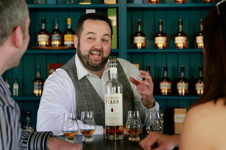 Man smiling and gesturing near whiskey bottle at bar with two people.