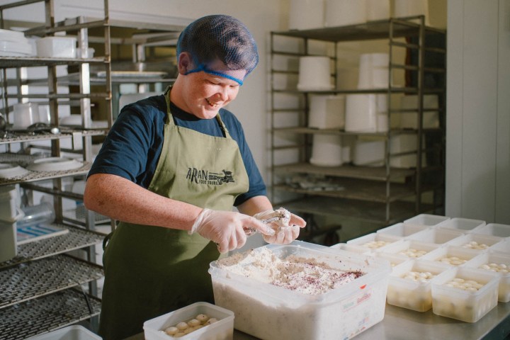 Person in apron and hairnet preparing food in a kitchen with shelves of containers.