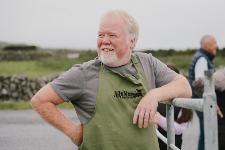 Smiling man in green apron stands by a fence in a rural setting.