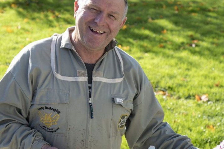 Smiling person in a beekeeping suit standing in a garden with flowers.