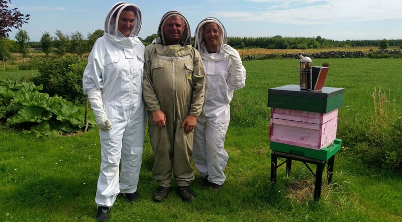Three beekeepers in suits stand next to a beehive in a grassy field.