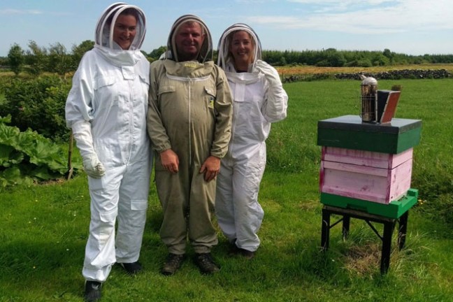 Three beekeepers in suits stand next to a beehive in a grassy field.