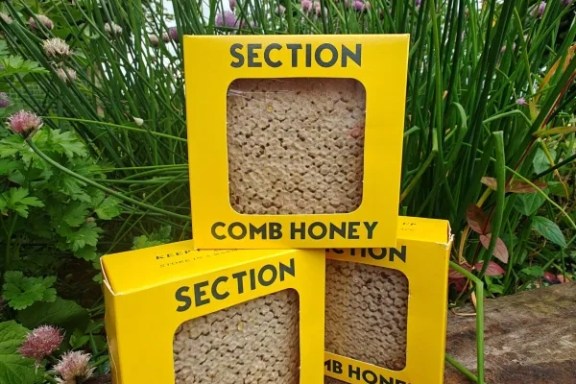 Three yellow boxes of comb honey on a wooden surface with purple flowers in the background.