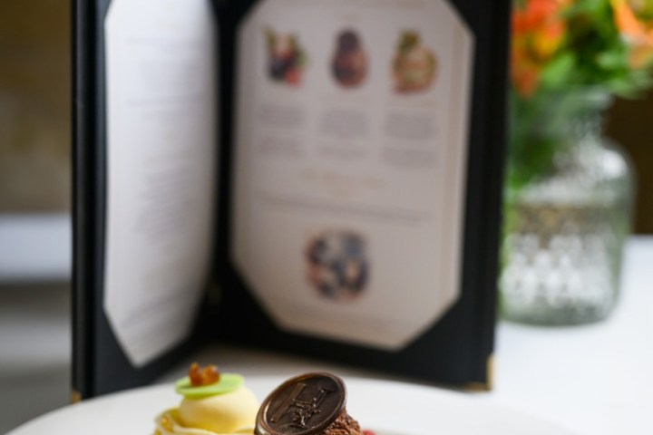 Elegant dessert trio on a plate with a blurred menu and flowers in the background.