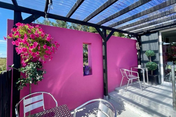 Outdoor seating area with pink wall, hanging flowers, and metal furniture under transparent roof.