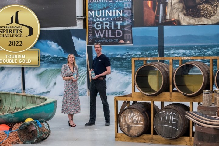 Two people hold bottles, standing by barrels and a boat. Award badge for 'International Spirits Challenge 2022' shown.