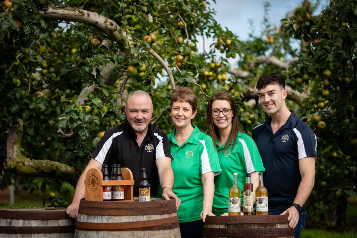 Four people standing by cider barrels in an apple orchard.