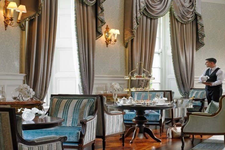 Elegant tearoom with waiter, draped curtains, chandeliers, and set tables.