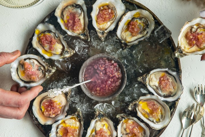 A platter of oysters with sauce on ice, surrounded by flowers and a glass of champagne.