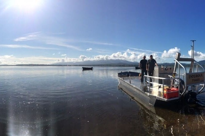 A boat on a calm lake, people on board, sunny sky, distant land and boats visible.