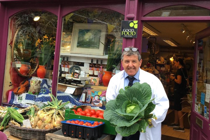 Man holding cabbage outside deli with fresh produce on display.